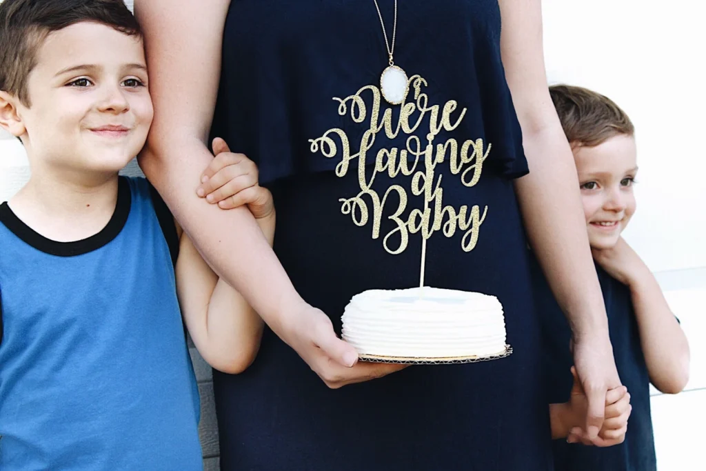 Pregnant mom holding hands of two sons while holding a white cake with a topper that says “we’re having a baby”.