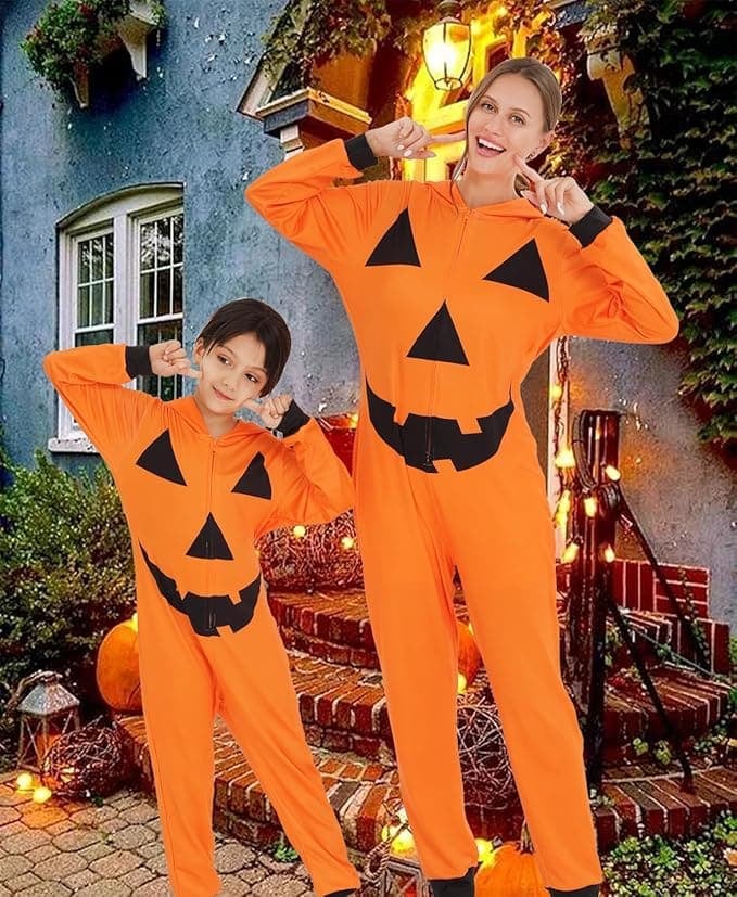 Mom and daughter wearing matching orange jack-o’-lantern jumpsuits posing playfully in front of a decorated Halloween porch with pumpkins and string lights