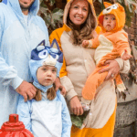 Family of four dressed in Bluey Halloween costumes — dad as Bandit, mom as Chilli, older daughter as Bluey, and baby as Bingo.