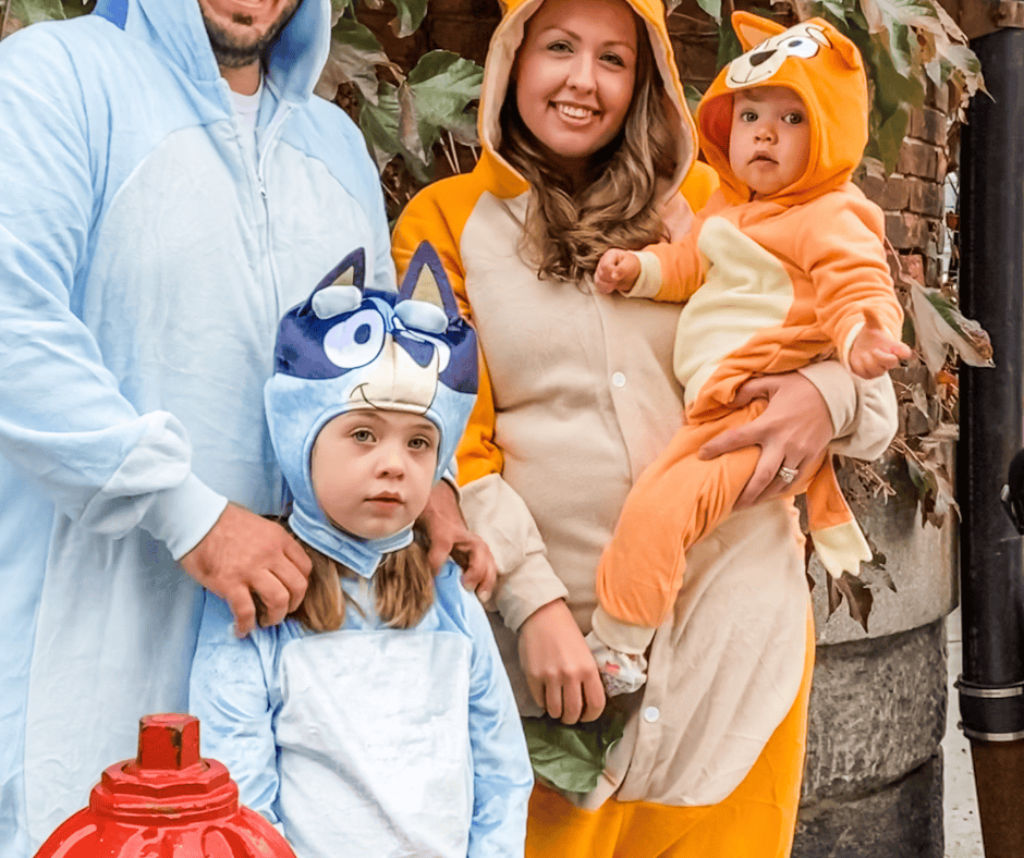 Family of four dressed in Bluey Halloween costumes — dad as Bandit, mom as Chilli, older daughter as Bluey, and baby as Bingo.