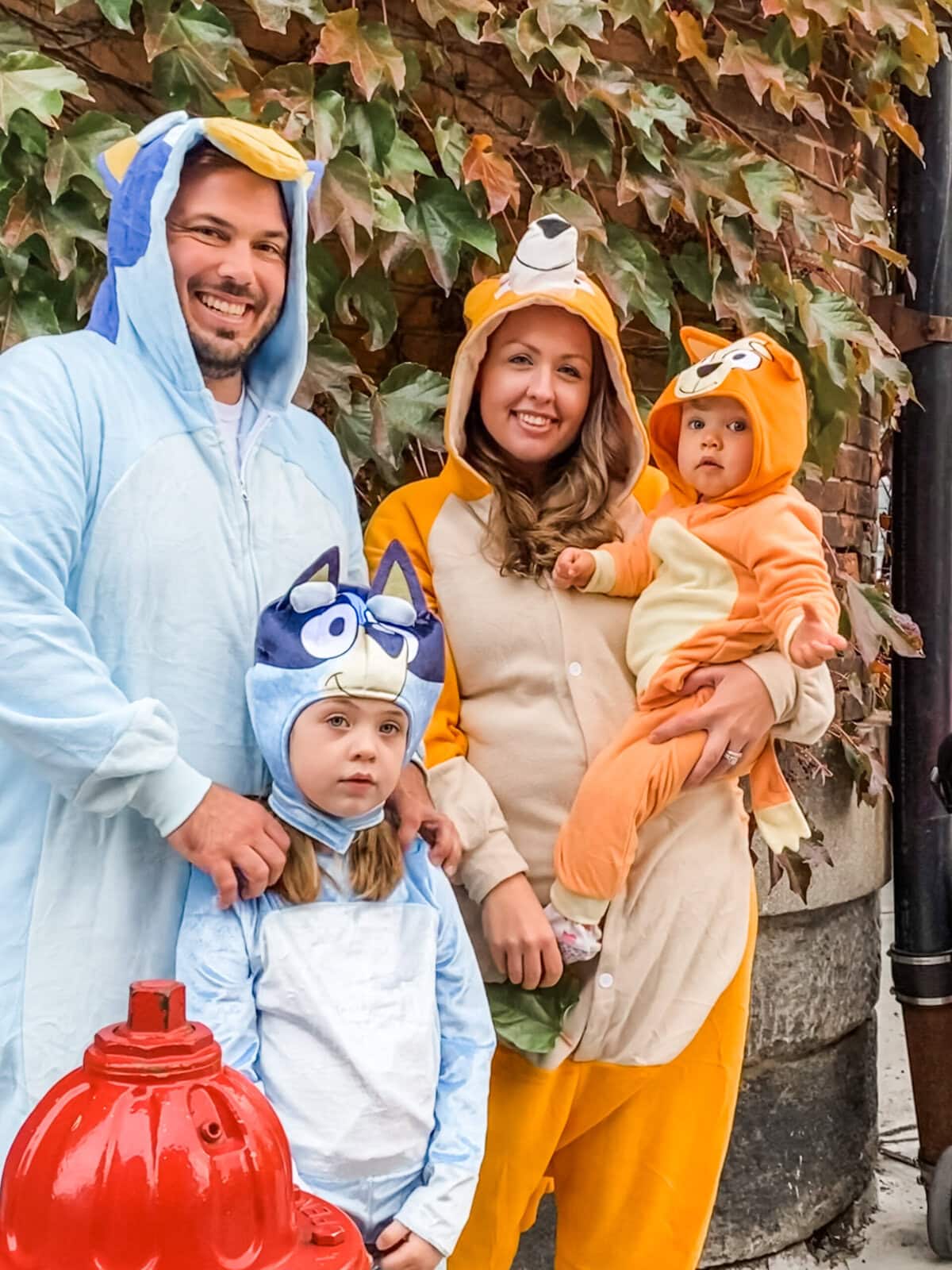 Family dressed in Bluey Halloween costumes posing outdoors — dad as Bandit, mom as Chilli, daughter as Bluey, and toddler as Bingo in cozy character onesies with fall leaves in the background