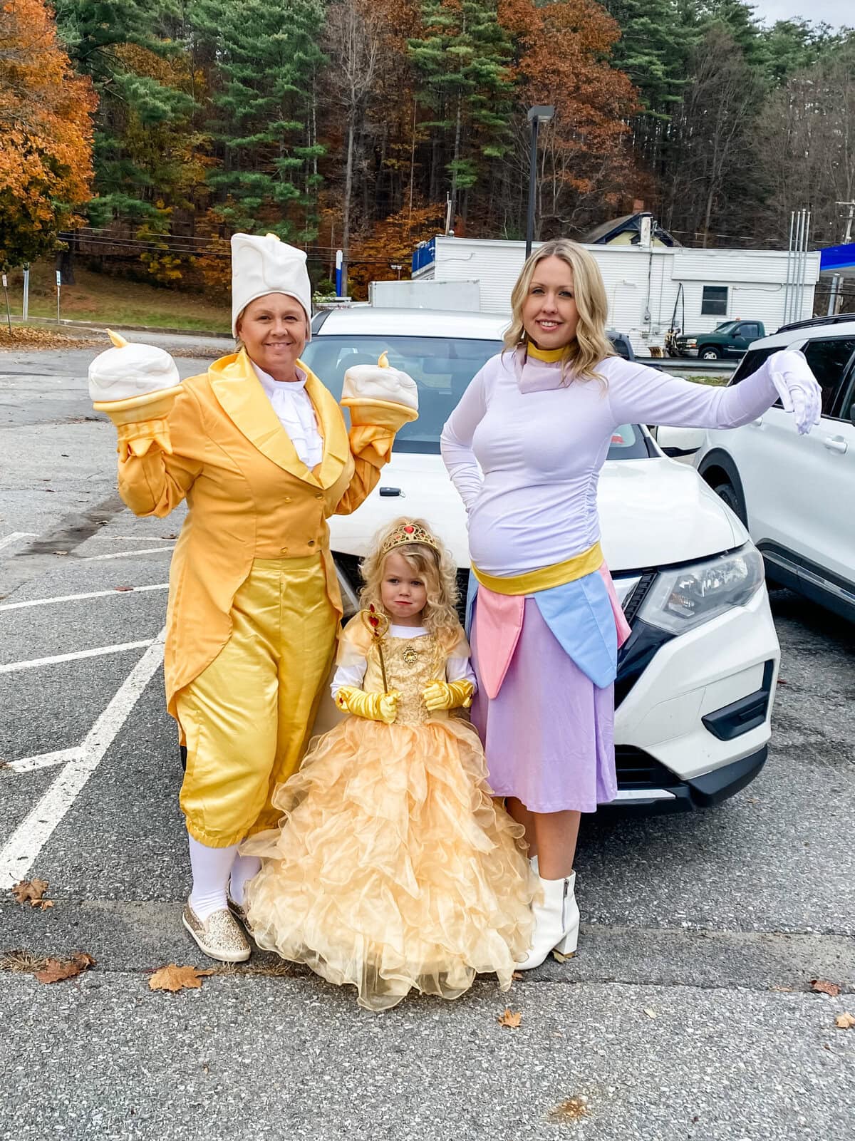 Family in Beauty and the Beast Halloween costumes standing in a parking lot with fall leaves — child dressed as Belle in a gold ball gown, adult as Lumière in a yellow suit with candle gloves, and pregnant mom in a DIY maternity Mrs. Potts costume with teapot-inspired skirt and white top.