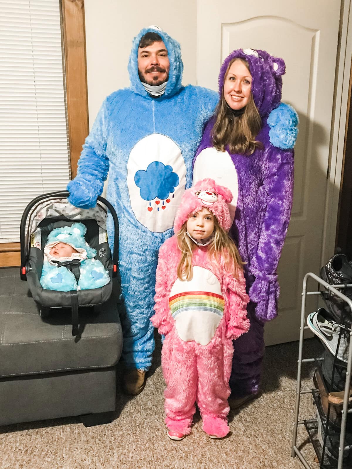 Family dressed in matching Care Bears Halloween costumes — dad in blue Grumpy Bear, mom in purple Share Bear, daughter in pink Cheer Bear, and baby in a light blue bear onesie in a car seat