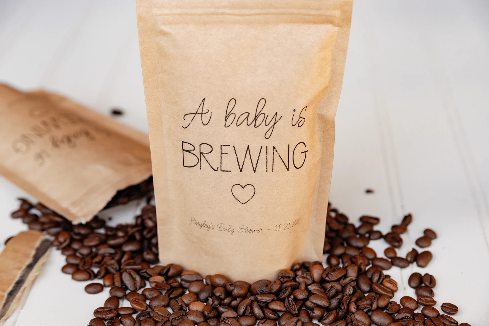“A baby is brewing” baby shower party favor coffee bags spilling coffee beans on a white table.