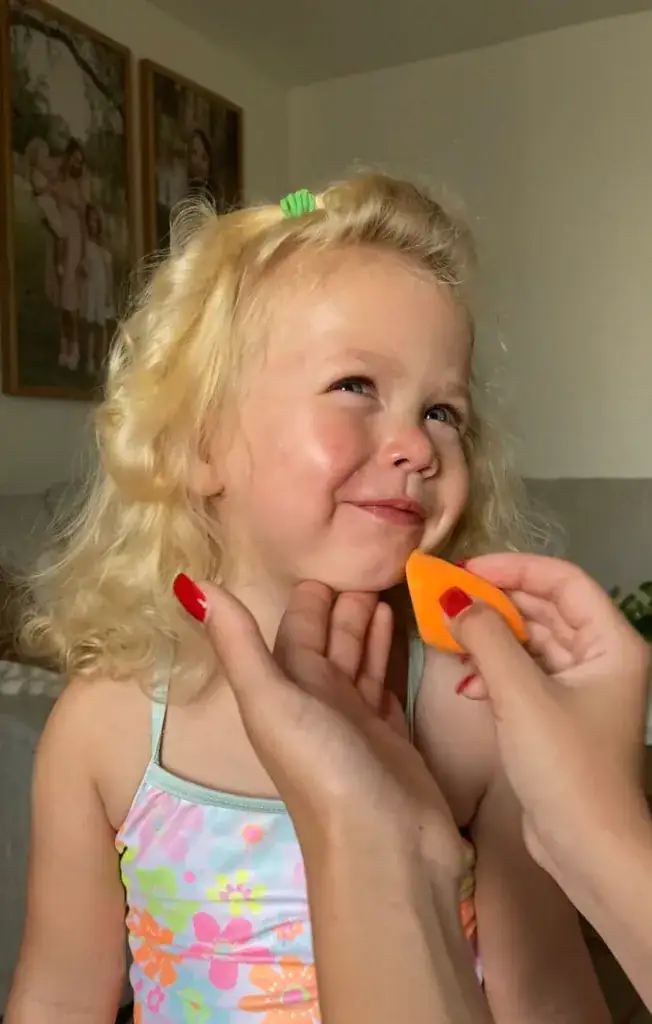 Young blonde girl in a floral swimsuit smiling as sunscreen is gently applied to her cheek with an orange makeup sponge.