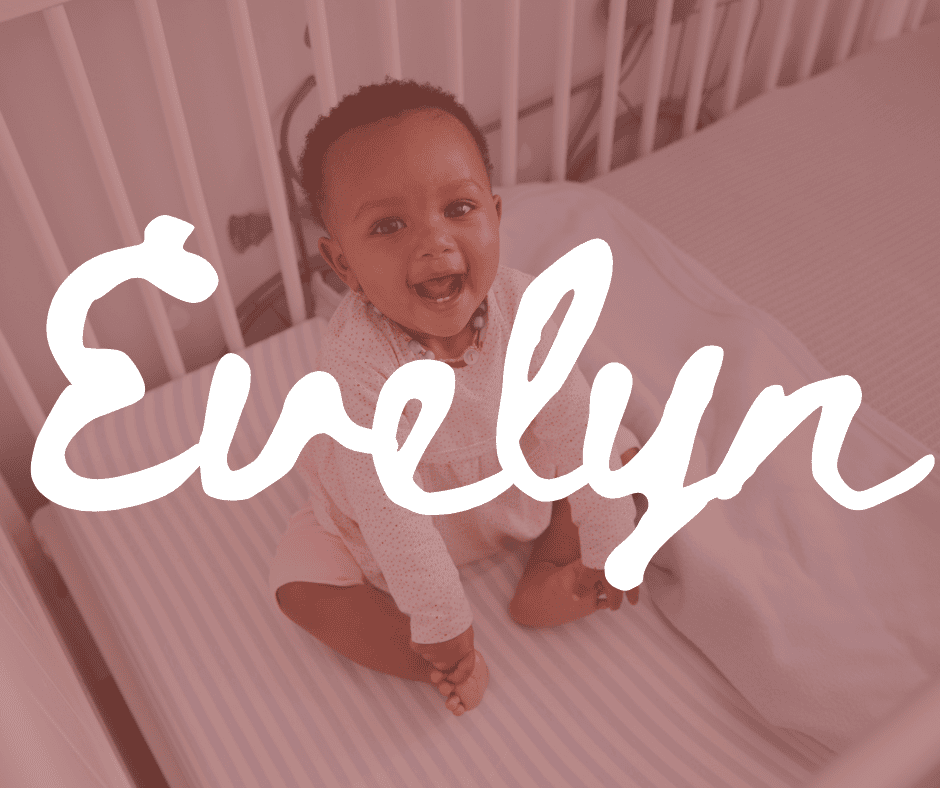 A happy smiling baby girl sits in a white crib. Overlay texts spells reads, “Evelyn”.