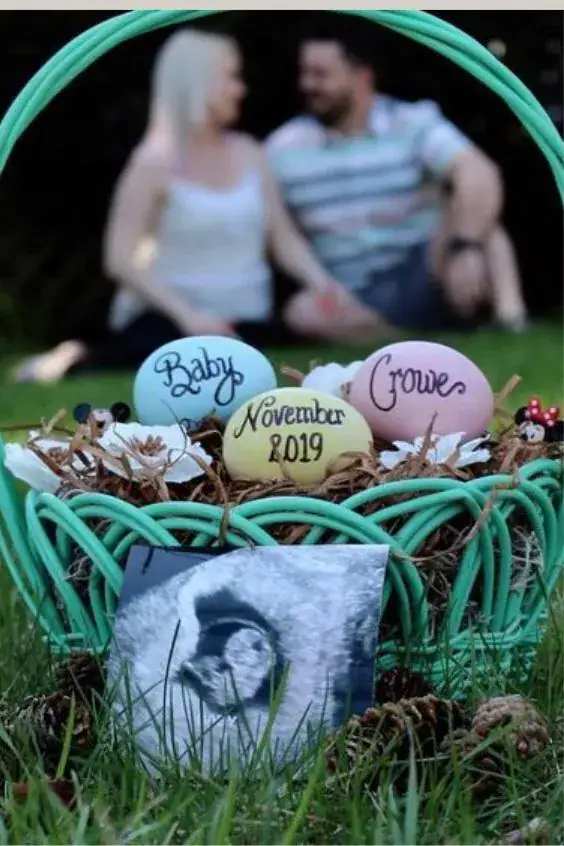 Easter pregnancy announcement photo. Three Easter eggs sit in an Easter basket. “Baby”, “Crowe” and “November 2019” are painted on the eggs. An ultrasound photo sits in front of the basket. The pregnant couple sits in the background blurred from vision.