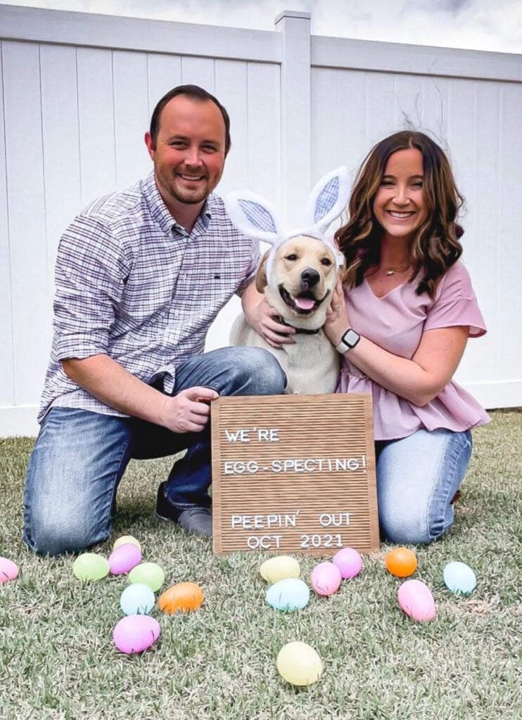 A smiling couple poses with their dog, who is wearing bunny ears, in a grassy yard with colorful Easter eggs scattered around. They hold a wooden letter board that reads, "We're EGG-specting! Peepin' out Oct 2021," announcing their pregnancy in a fun Easter-themed way.
