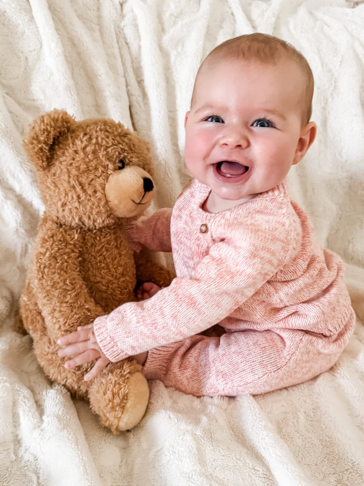 Six-month-old baby girl sitting up in a knit pink outfit, laughing and reaching for her teddy bear during a monthly milestone photo at home.