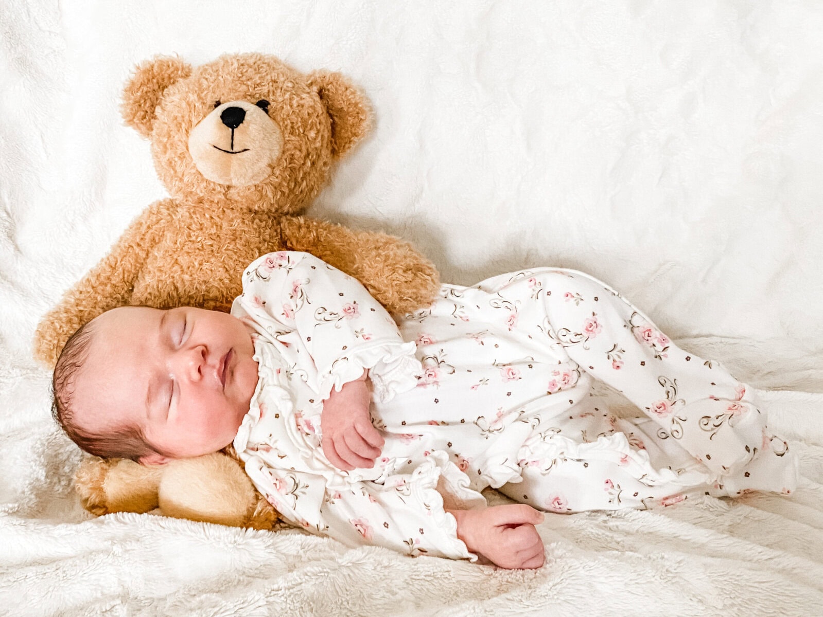 Newborn baby girl sleeping in a floral sleeper, nestled against a brown teddy bear on a white fuzzy blanket for a cozy at-home milestone photo.