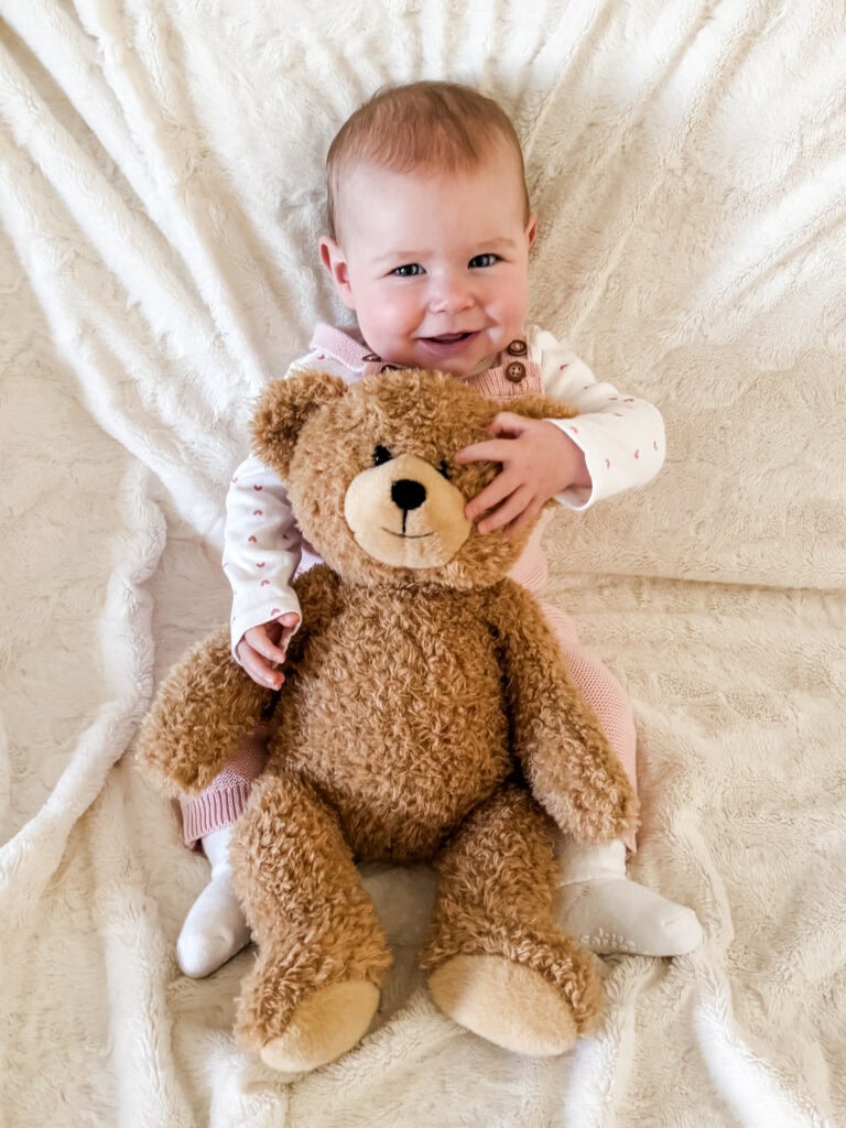 Seven-month-old baby in pink overalls sitting behind a teddy bear, reaching around and playfully grabbing its face for her milestone photo.