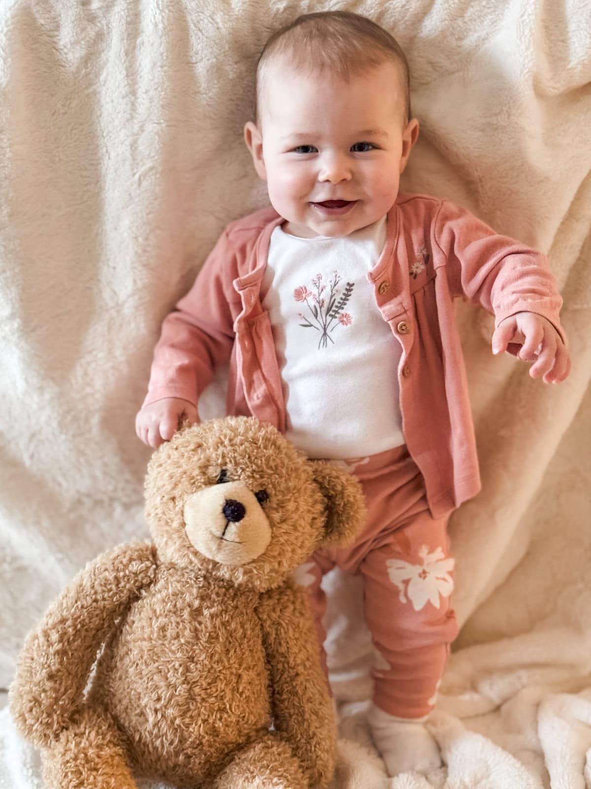 Eight-month-old baby standing with support against a couch, wearing a pink floral outfit and smiling beside her seated teddy bear.
