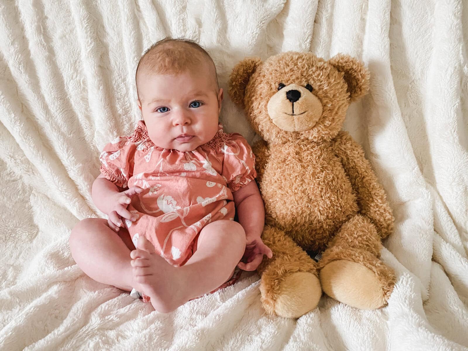 Three-month-old baby dressed in a coral floral romper sitting next to a teddy bear on a white blanket for a monthly milestone photo at home.