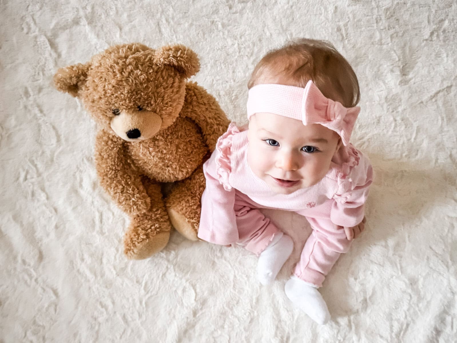 Nine-month-old baby girl dressed in pink with a big bow, sitting on a white blanket next to her teddy bear in an overhead photo taken at home.