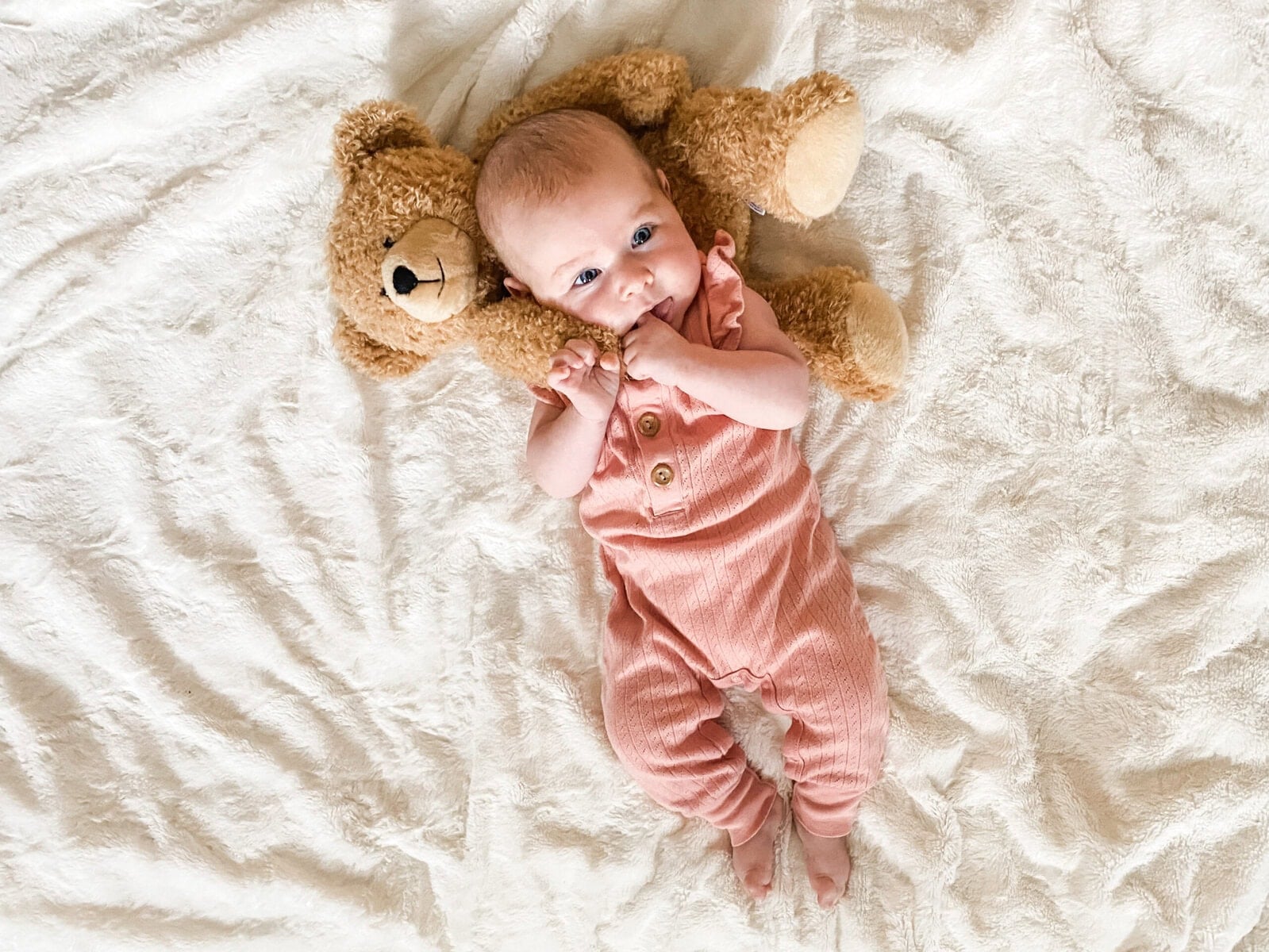Four-month-old baby girl lying on a white blanket in a ribbed pink romper, sucking on her hands with a teddy bear behind her head.
