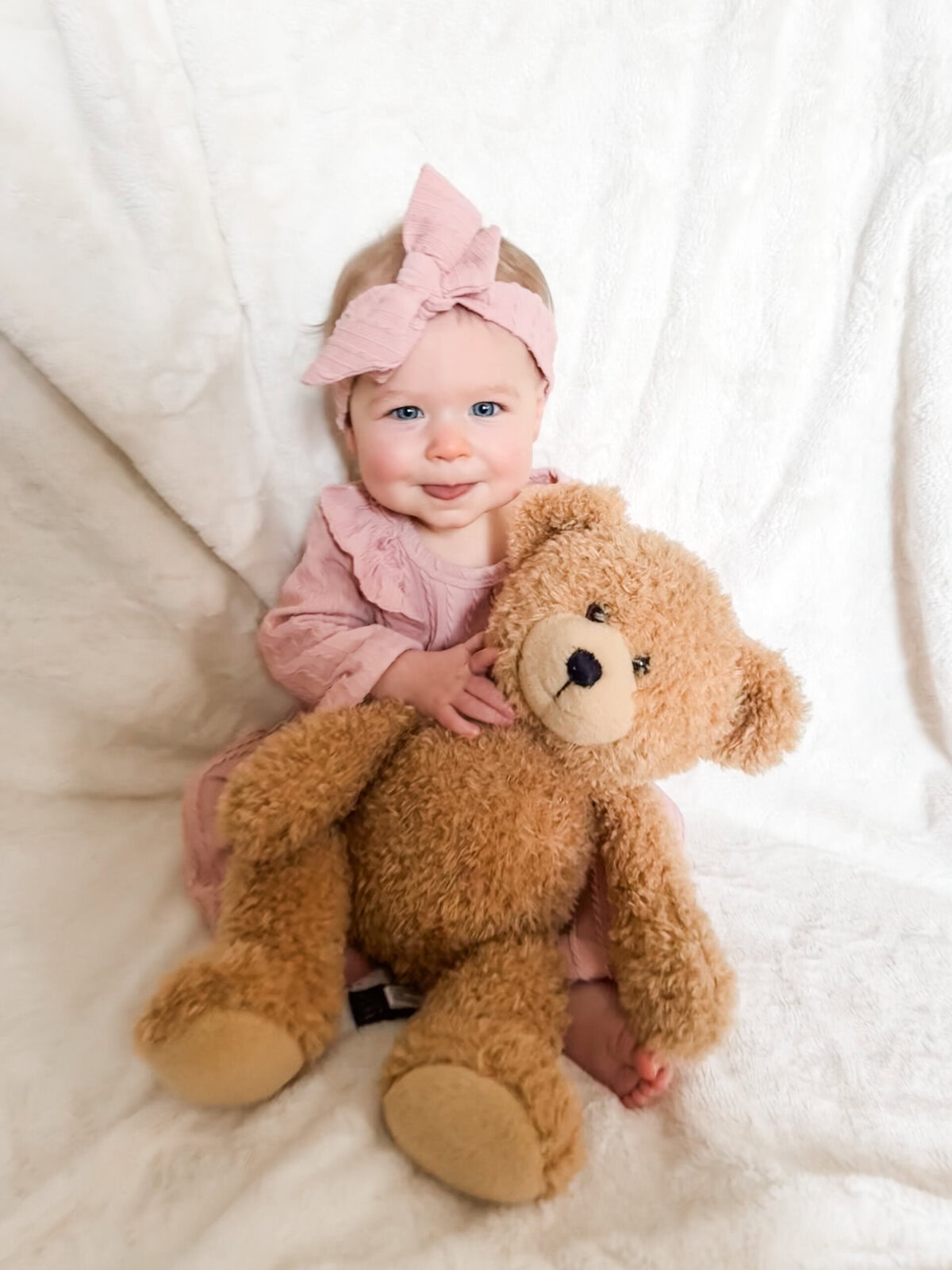 Ten-month-old baby in a dusty rose ruffled outfit, sitting upright and gently holding her teddy bear while glancing at the camera.