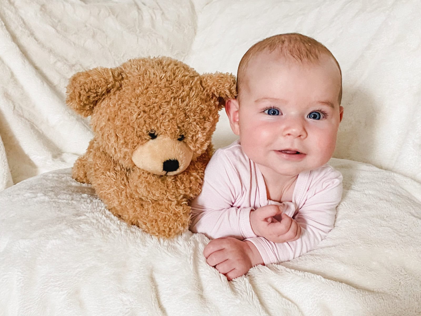 Five-month-old baby in a light pink onesie doing tummy time on a couch with a white blanket, smiling beside her teddy bear.
