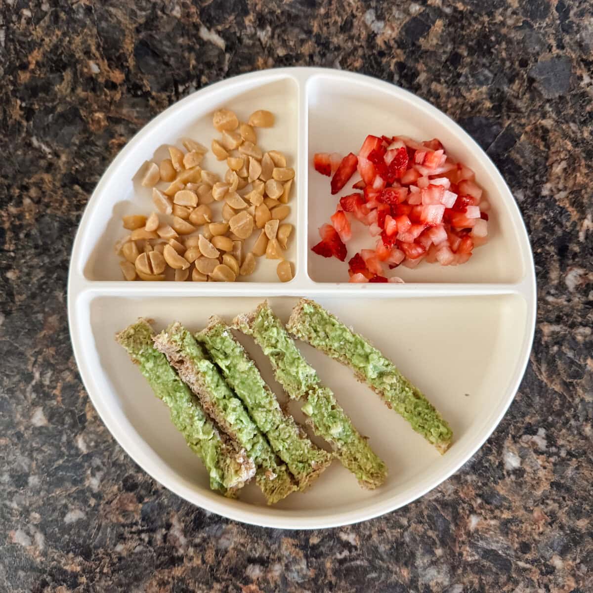 A toddler lunch plate with strips of avocado toast on Ezekiel bread, sliced chickpeas, and diced strawberries on a cream-colored divider plate.