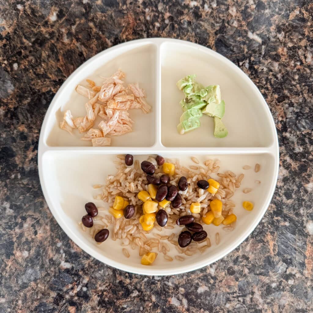 A toddler lunch plate with brown rice topped with black beans and corn, shredded chicken, and diced avocado on a cream-colored divider plate.