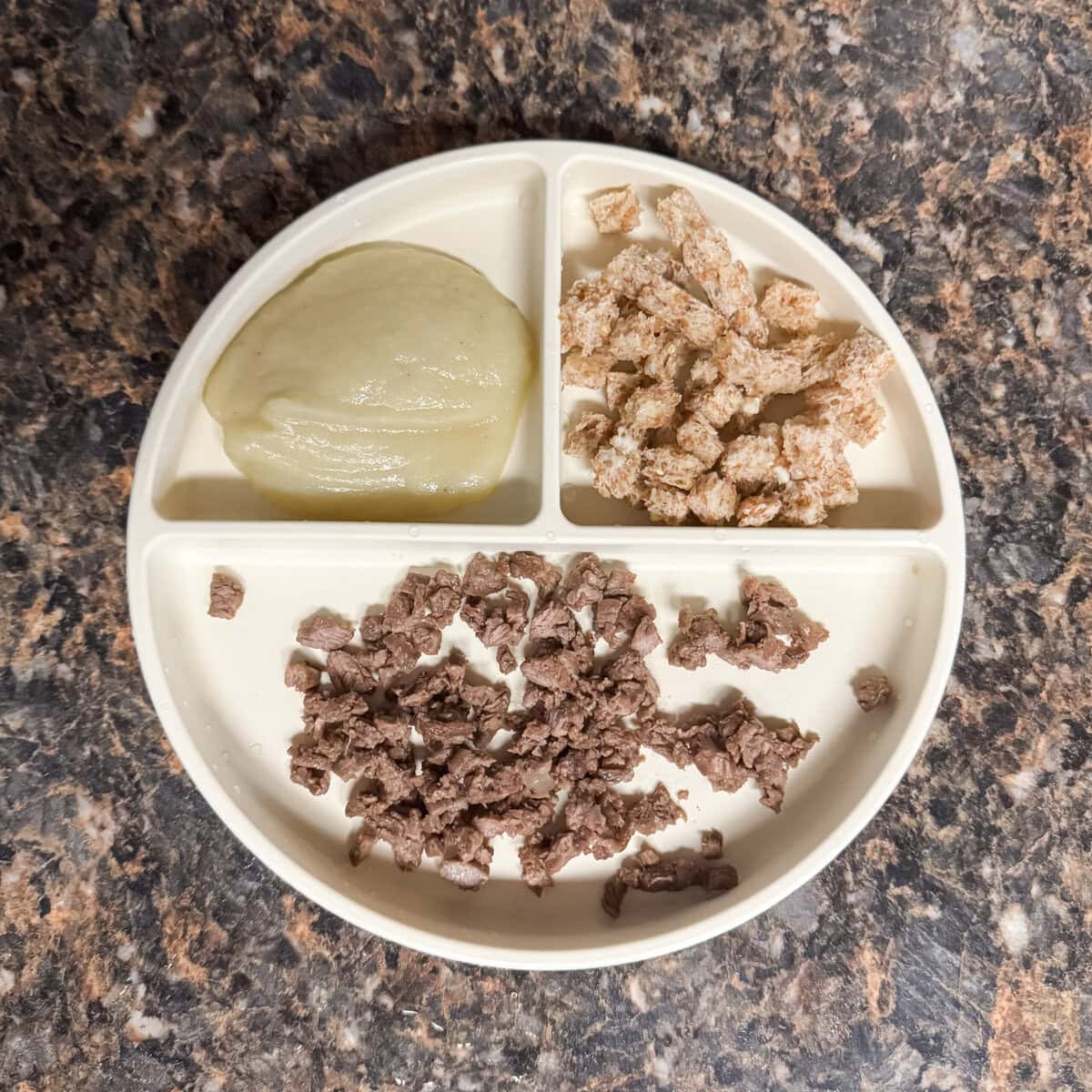 A toddler lunch plate with chopped carne asada steak, mashed cauliflower, and torn buttered Ezekiel toast on a cream-colored divider plate.