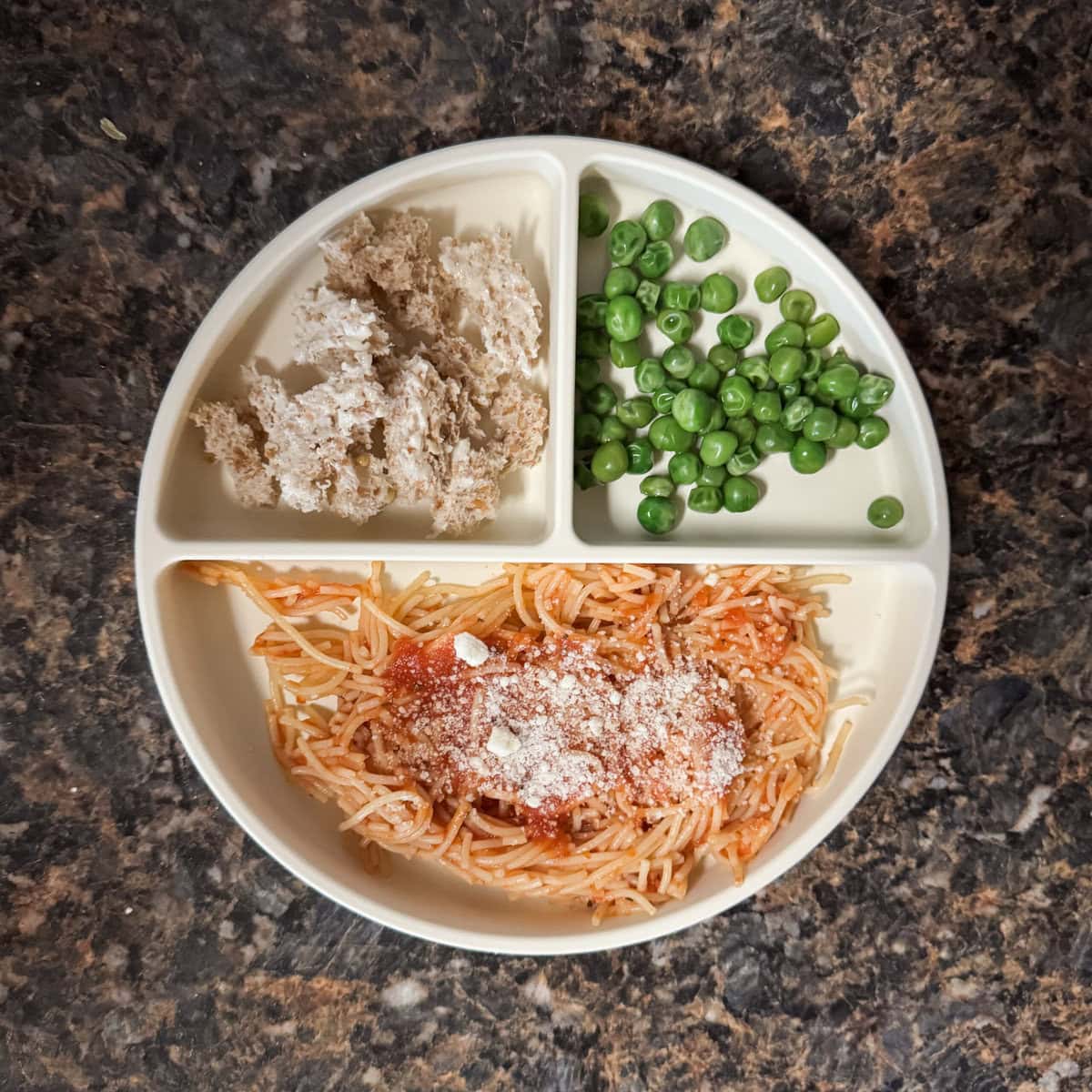 A toddler lunch plate with spaghetti topped with marinara and Parmesan, steamed peas, and torn pieces of buttered Ezekiel toast on a cream-colored divider plate.