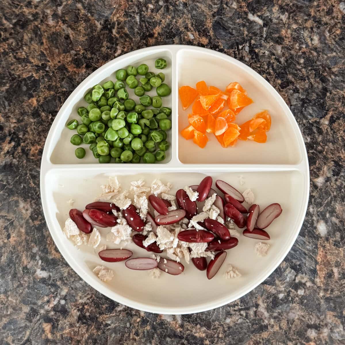 A toddler lunch plate with shredded chicken and halved kidney beans, peeled clementine segments, and steamed green peas on a cream-colored divider plate.