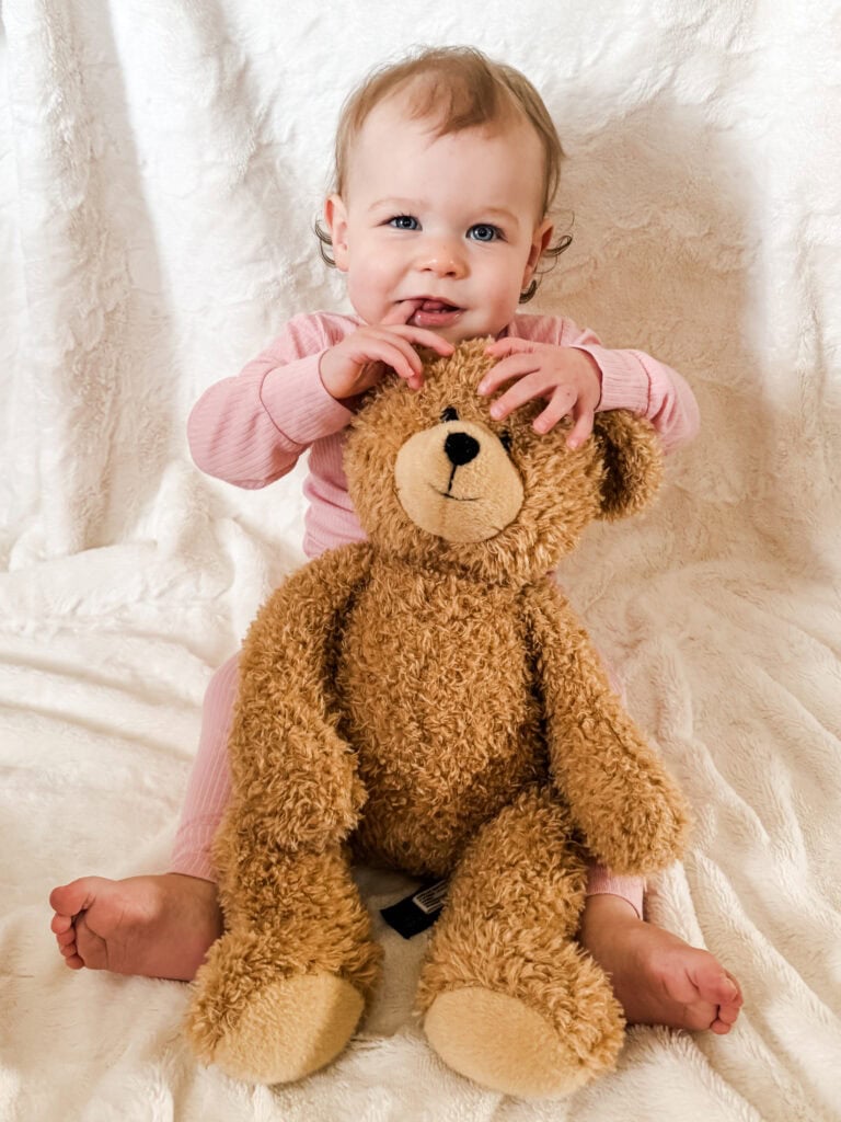 One-year-old baby girl sitting on a white blanket in a pink outfit, holding her teddy bear in front of her with a sweet expression.