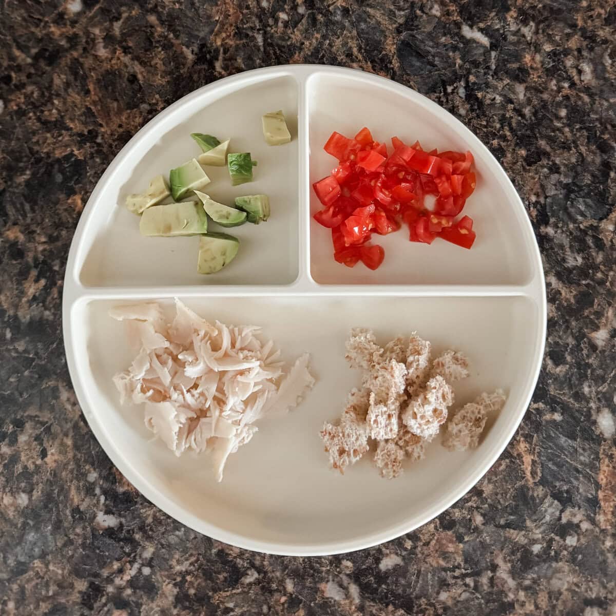A toddler lunch plate with shredded deli turkey, torn pieces of Ezekiel flourless bread, diced tomato, and diced avocado on a cream-colored divider plate.
