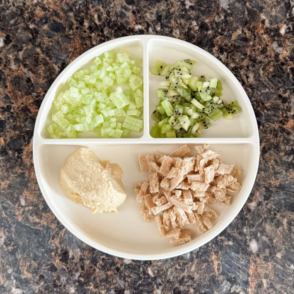 A toddler lunch plate with chopped whole wheat pita, a scoop of hummus, diced cucumber, and diced kiwi on a cream-colored divider plate.