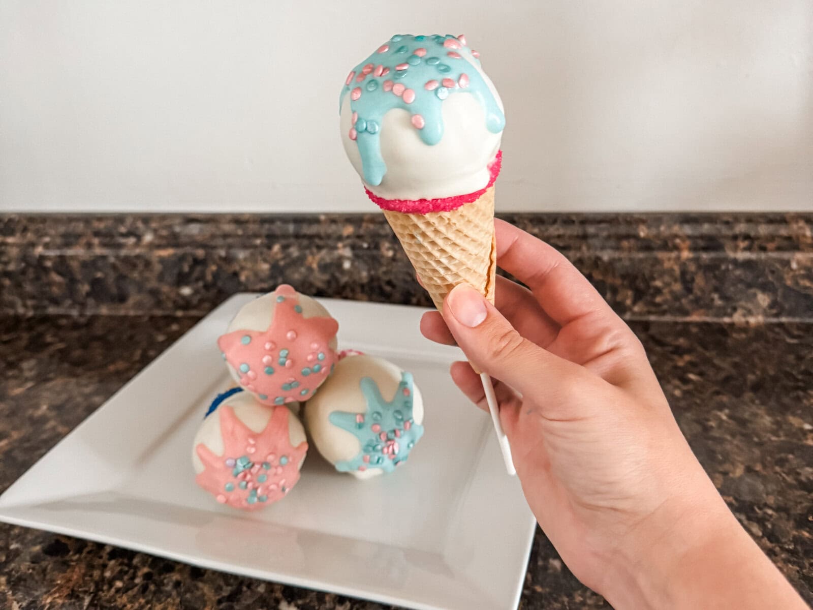 Close-up of a finished ice cream cone gender reveal cake pop decorated with blue drizzle and pink sprinkles. In the background, more cake pops with pink and blue drizzle rest on a white square plate atop a brown granite countertop.