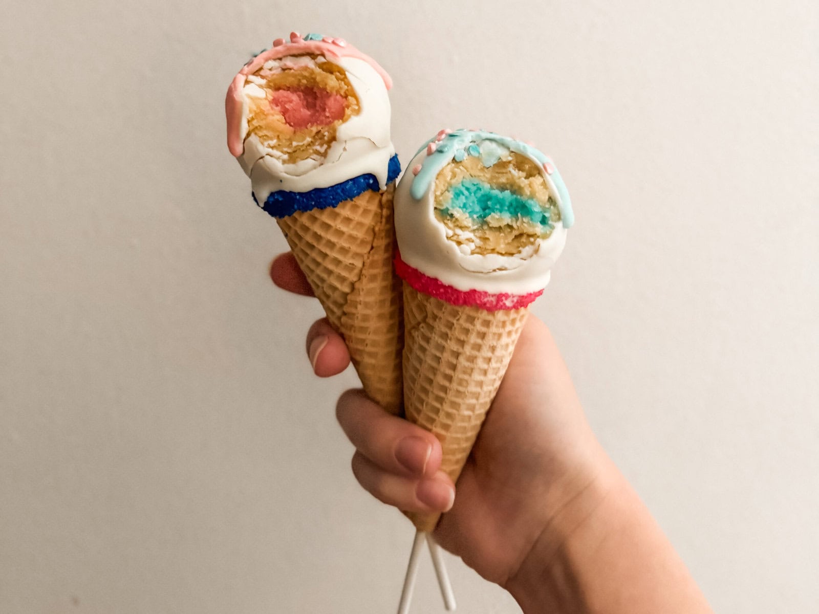 Hand holding two ice cream cone gender reveal cake pops with a bite taken out of each. One reveals a pink cake center and the other reveals a blue cake center. Both are dipped in white chocolate and decorated with colorful drizzle and sprinkles.