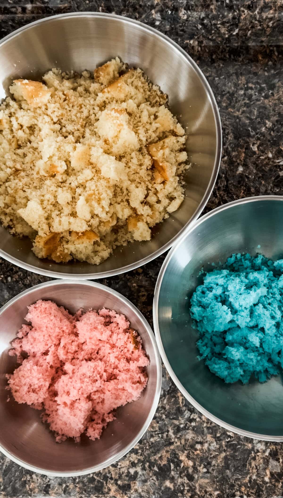 Overhead view of three stainless steel mixing bowls filled with crumbled cake on a dark brown granite countertop. One bowl contains plain white cake, while the other two hold cake tinted pink and blue, prepared for gender reveal cake pops.