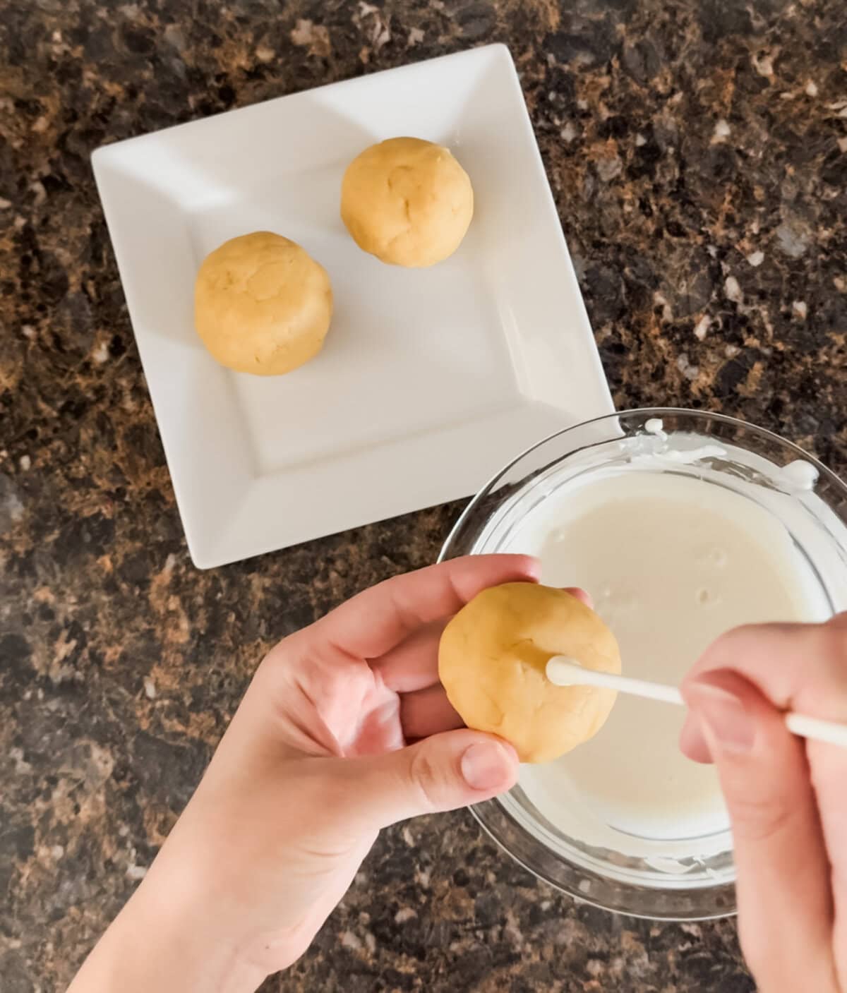 Close-up of hands dipping a white cake pop stick into melted white chocolate before inserting it into a smooth cake ball. Two additional cake balls rest on a square white plate in the background, all set on a dark granite countertop.