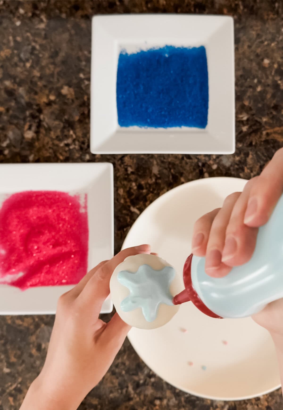 Close-up of hands decorating a gender reveal cake pop with blue chocolate drizzle using a squeeze bottle. Plates of pink and blue sanding sugar are visible in the background, set on a dark granite countertop.