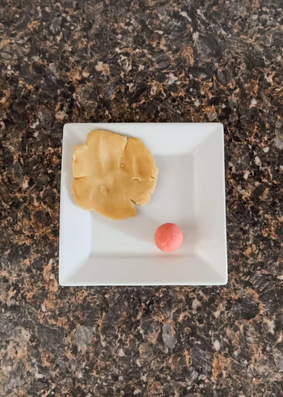 Over head view of a flat circle of white cake pop dough and small pink sphere of cake pop dough on a white plate placed on a dark granite countertop.
