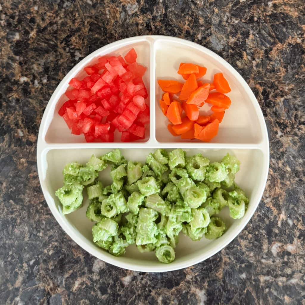 A toddler lunch plate with pasta coated in a green pea and Parmesan sauce, diced watermelon, and steamed carrot slices served in a cream-colored divider plate.
