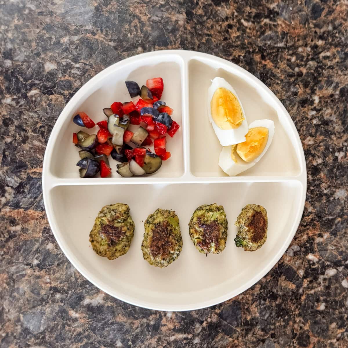A toddler lunch plate with four broccoli cheddar tots, two quartered slices of a hard-boiled egg, and a side of chopped strawberries and blueberries on a cream-colored divider plate.