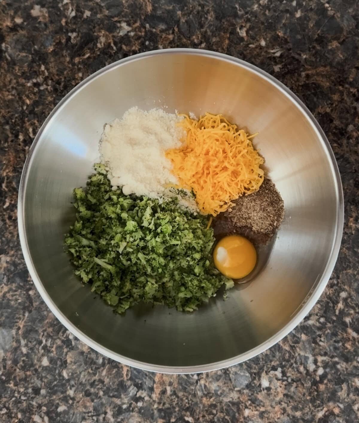 Overhead view of a stainless steel mixing bowl sitting on a dark marbled countertop. Inside the bowl are neatly separated ingredients for broccoli cheddar tots: finely chopped steamed broccoli, shredded cheddar cheese, grated Parmesan cheese, ground flaxseed, and a whole egg with the yolk intact. Each ingredient is placed in its own section, showing a colorful and organized layout before mixing.