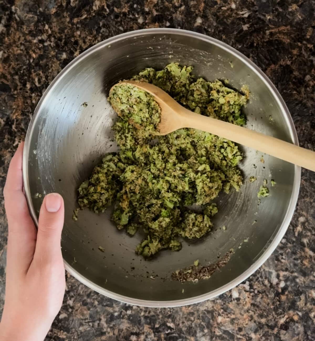 A hand holds a stainless steel mixing bowl filled with a blended broccoli cheddar tot mixture. The ingredients have been fully combined, forming a thick green mixture with visible bits of broccoli. A wooden spoon rests in the bowl, coated in the mixture, against a marbled dark countertop background.