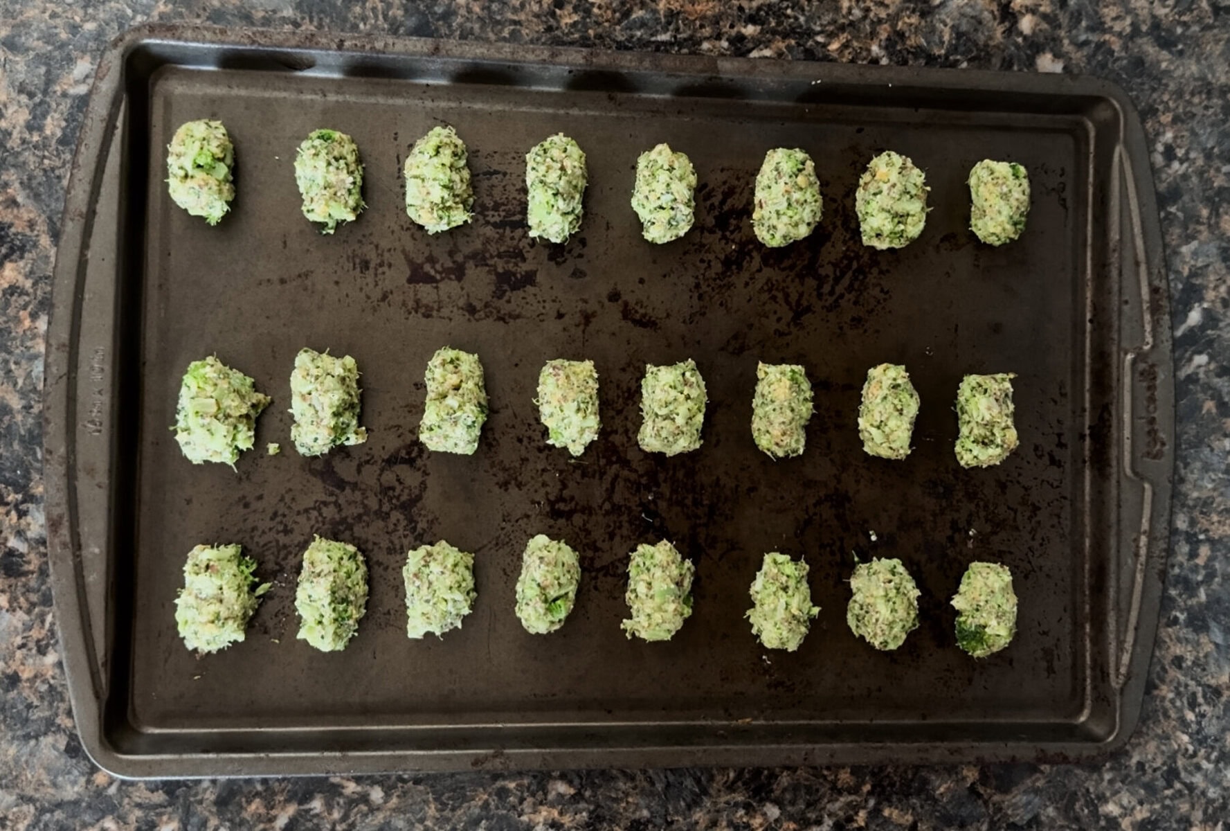 A baking sheet with twenty-four unbaked broccoli cheddar tots neatly arranged in rows. Each tot is shaped into a small oval and evenly spaced on the dark, slightly worn baking tray. The vibrant green flecks of broccoli are visible in the mixture, resting on a marbled dark countertop.