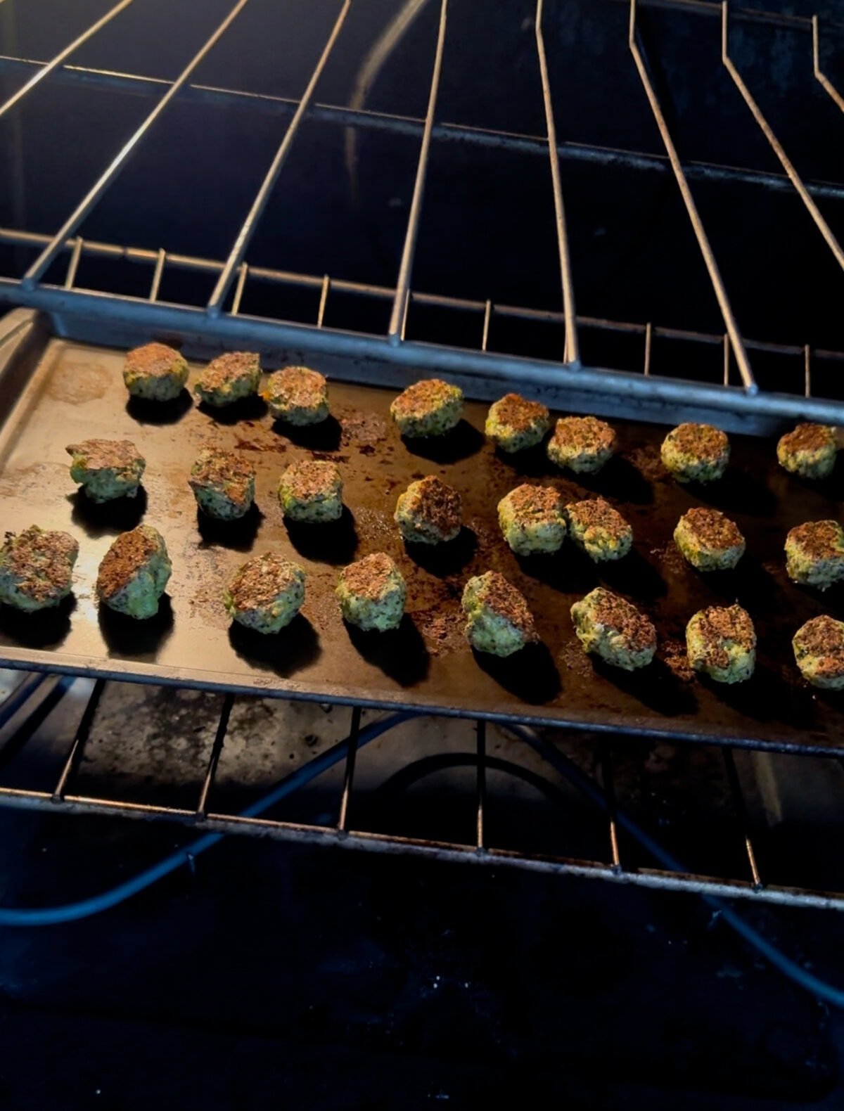 A tray of broccoli cheddar tots baking in the oven, captured mid-cook. The tots are golden brown on top and arranged in even rows on a dark baking sheet, set on the middle rack of a lit oven. The warm glow highlights their crisped tops and rustic texture.