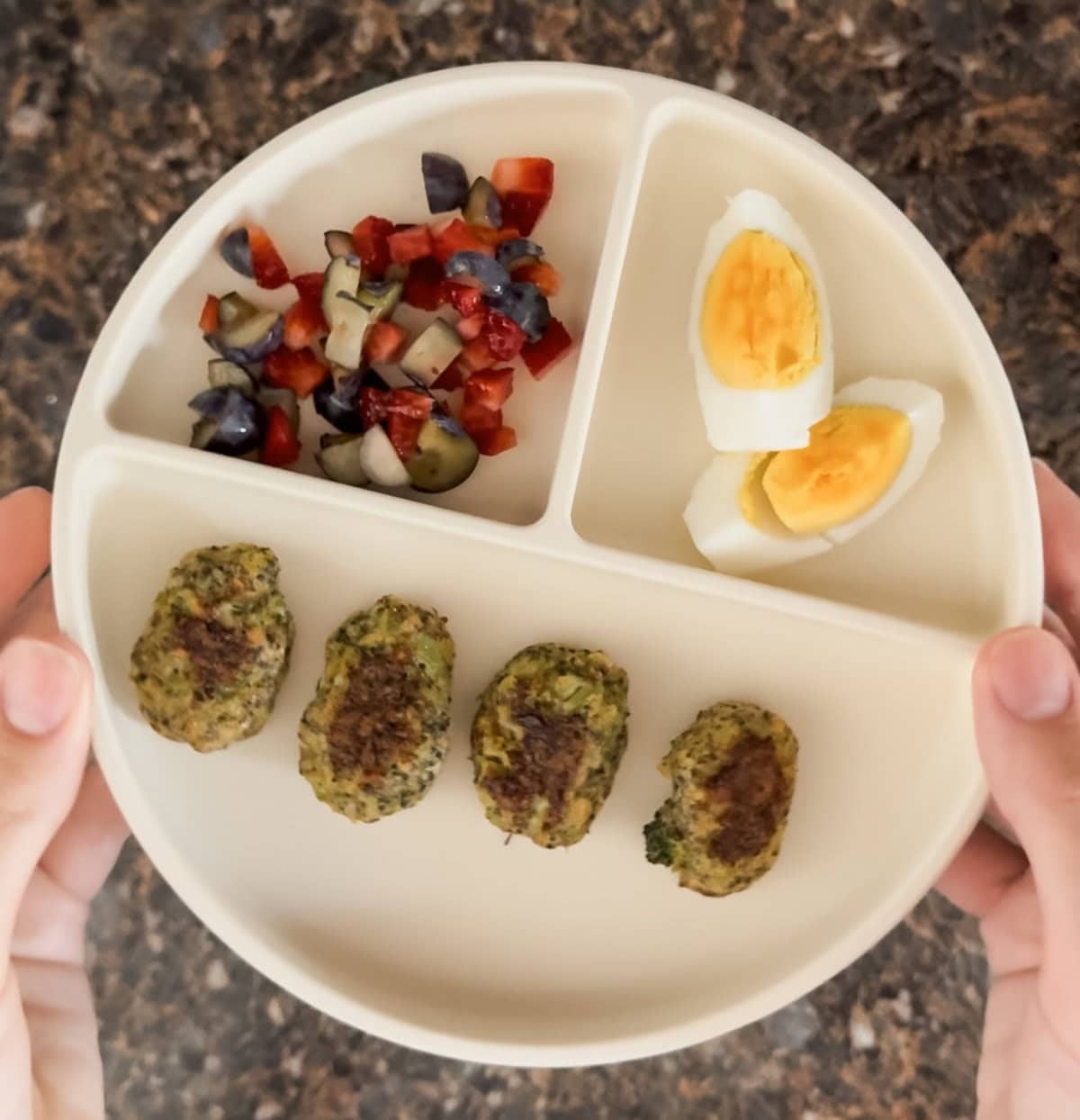 A beige toddler plate with three sections, held above a granite countertop. One section contains four golden-brown broccoli cheddar tots, the second has a colorful mix of chopped blueberries and strawberries, and the third holds a sliced hard-boiled egg. The meal is vibrant, balanced, and perfect for toddlers.