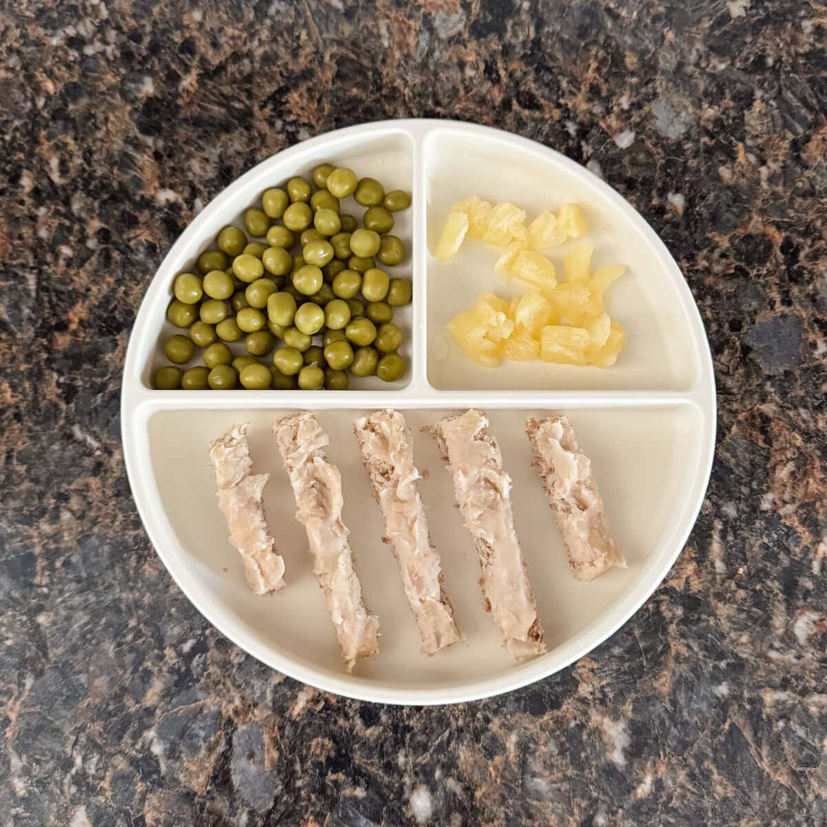 A toddler lunch plate with strips of Ezekiel toast topped with mashed white kidney beans, chopped pineapple, and green peas on a cream-colored divider plate.