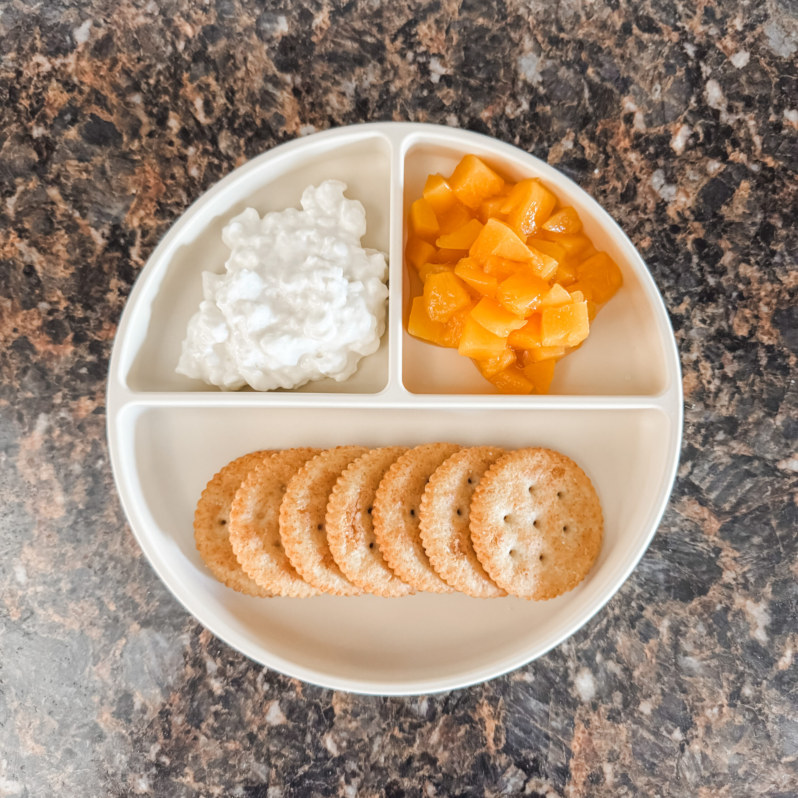 A divided white toddler plate on a marble countertop filled with round crackers in the bottom section, a scoop of cottage cheese in the left section, and diced peaches in the right section.
