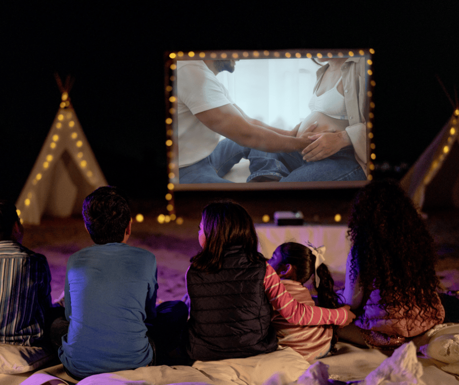 Guests gather around an outdoor movie screen at a summer gender reveal party. A gender reveal video plays on the screen.