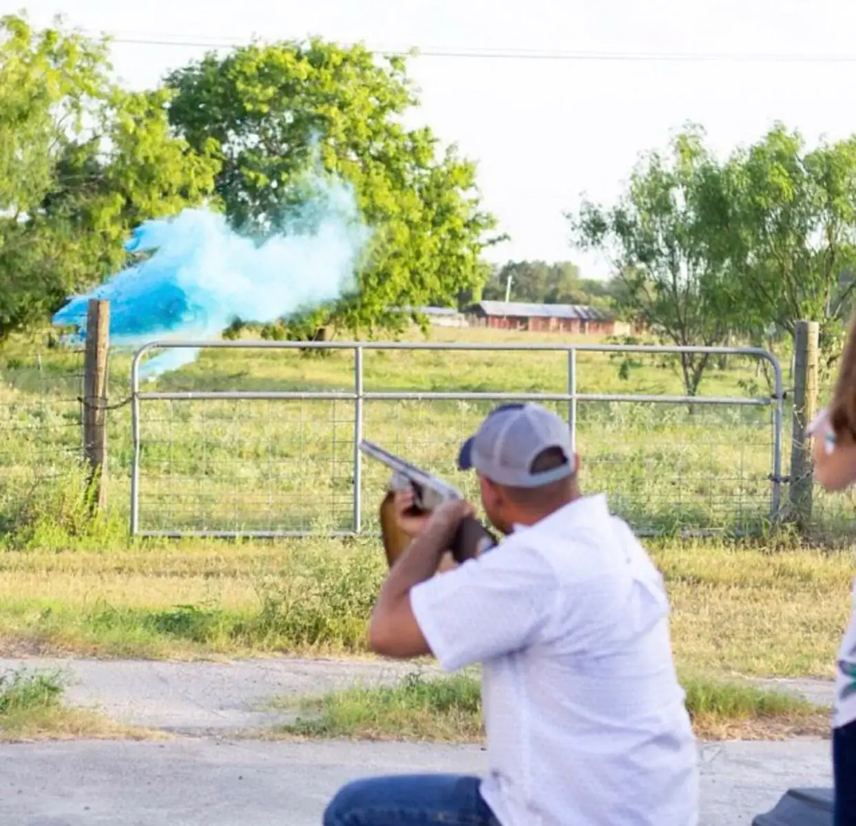 A father shoots at a target that explodes with blue smoke at a summer gender reveal party.