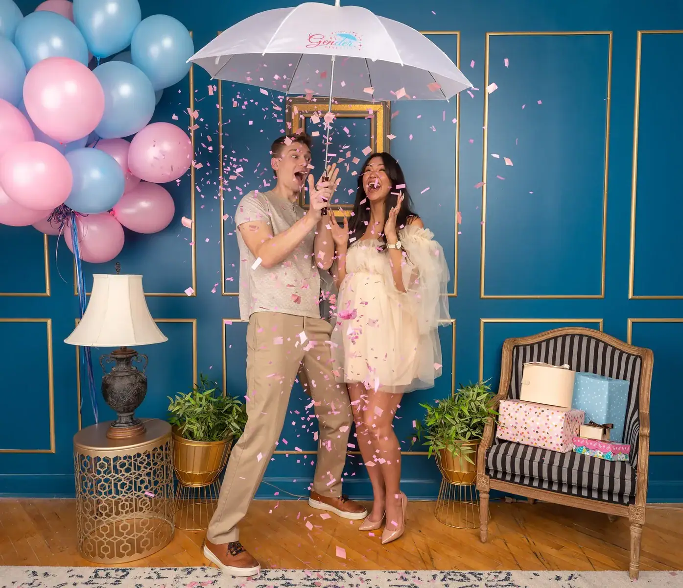 A pregnant couple opens an umbrella over their heads and pink confetti falls.