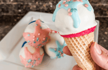 Close-up of a hand holding a gender reveal cake pop styled like an ice cream cone, with blue drizzle and pink and blue sprinkles on top of white chocolate. Three additional cake pops with pink and blue drizzles are stacked on a white plate in the background.