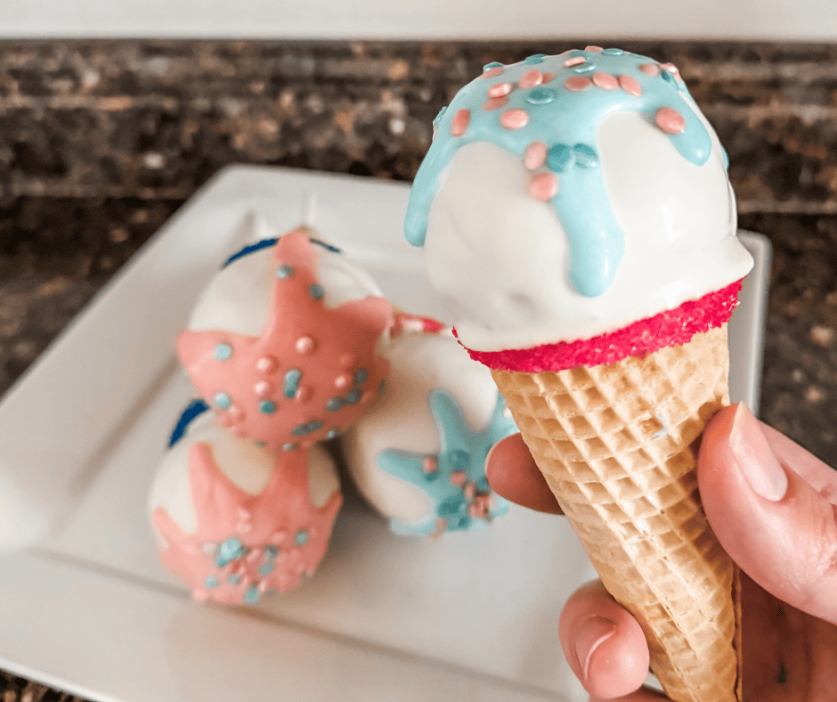 Close-up of a hand holding a gender reveal cake pop styled like an ice cream cone, with blue drizzle and pink and blue sprinkles on top of white chocolate. Three additional cake pops with pink and blue drizzles are stacked on a white plate in the background.