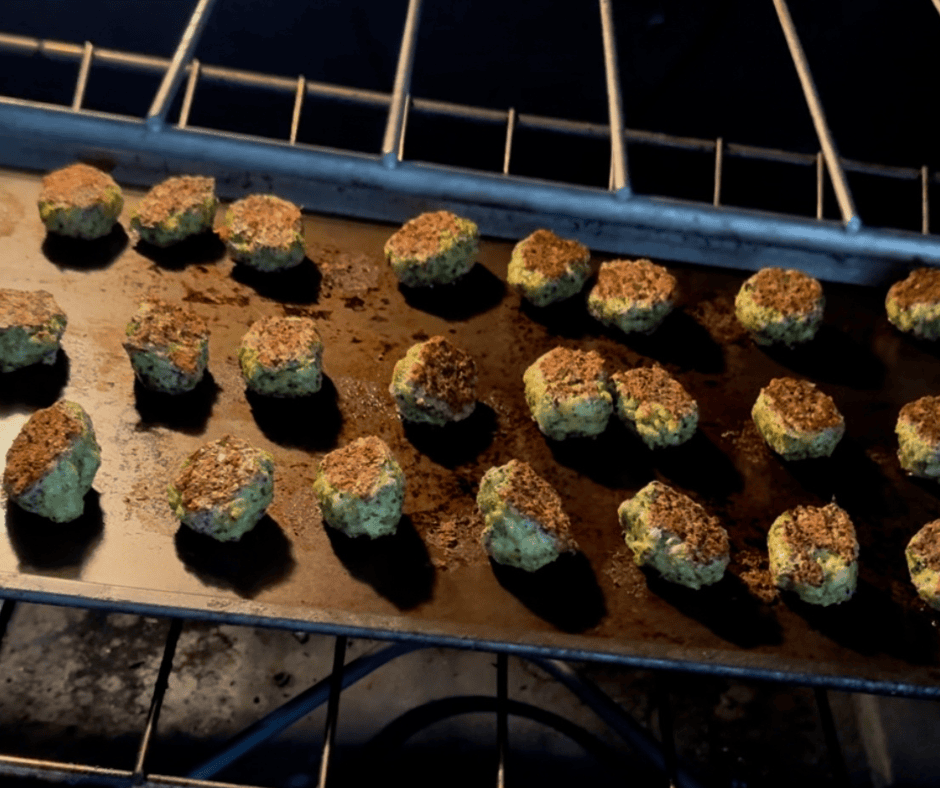A tray of broccoli cheddar tots baking in the oven, captured mid-cook. The tots are golden brown on top and arranged in even rows on a dark baking sheet, set on the middle rack of a lit oven. The warm glow highlights their crisped tops and rustic texture.